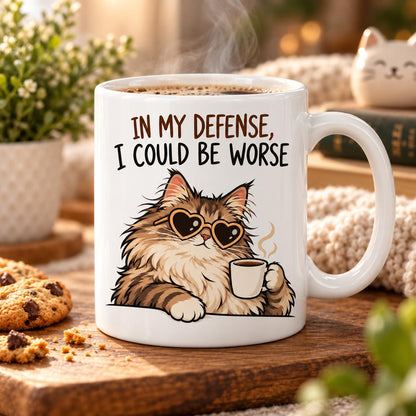 Rendering of white ceramic mug with cartoon content maincoon cat saying "In my defence, I could by worse" on wooden table. 