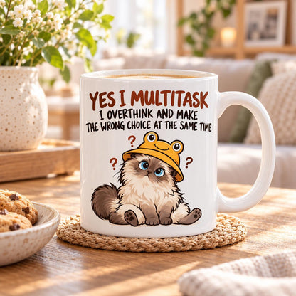 Rendering of white ceramic mug with cartoon confused ragdoll cat saying "Yes I can multitask. I overthing ane make the wrong choice" on wooden table.