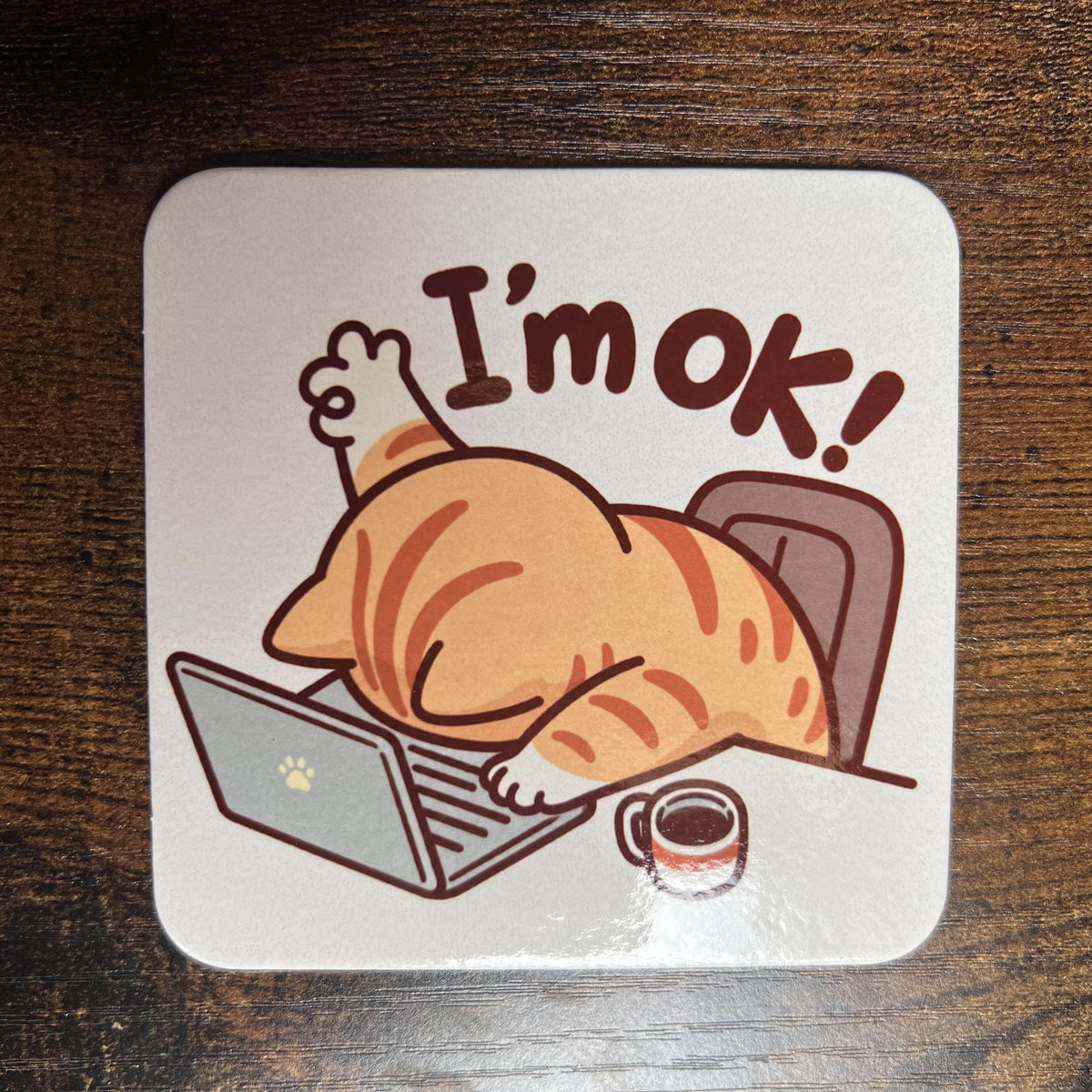 Korkový podtácek s unavenou kreslenou kočkou ukazující "I'm OK" na dřevěném stole. Pohled shora.