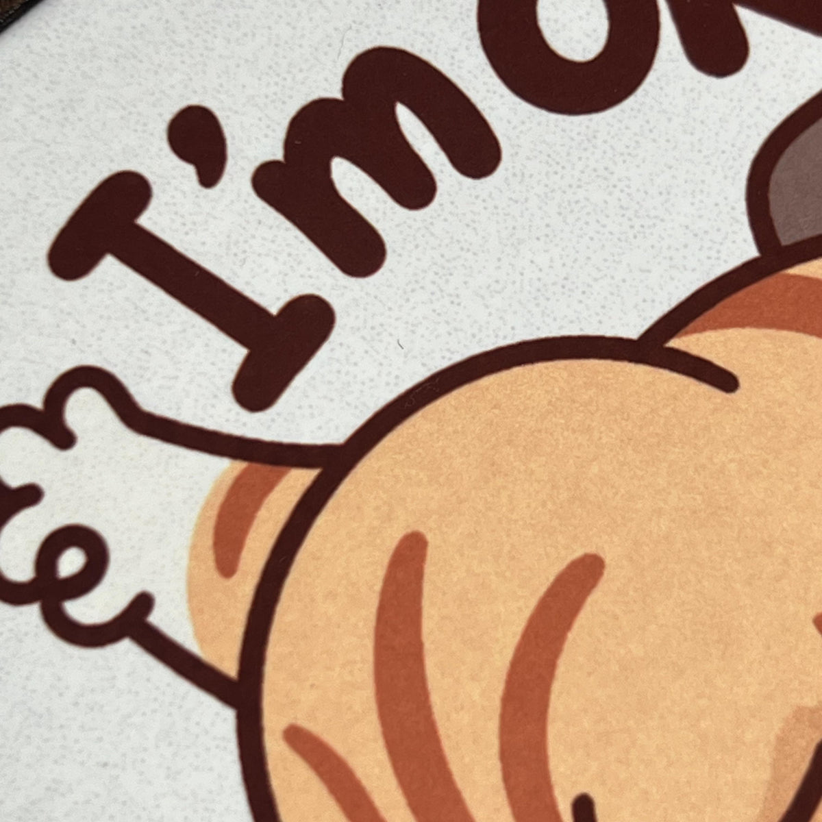Detail tisku na korkovém podtácku s unavenou kreslenou kočkou ukazující "I'm OK".