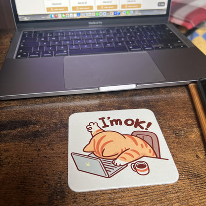 Korkový podtácek s unavenou kreslenou kočkou ukazující "I'm OK" na dřevěném stole před počítačem. 