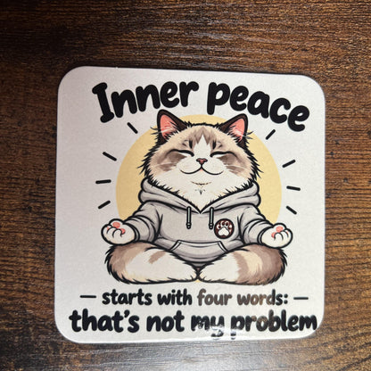 Korkový podtácek s kreslenou meditující ragdoll kočkou říkající "Inner peace starts with four words ..." na dřevěném stole. Pohled shora.