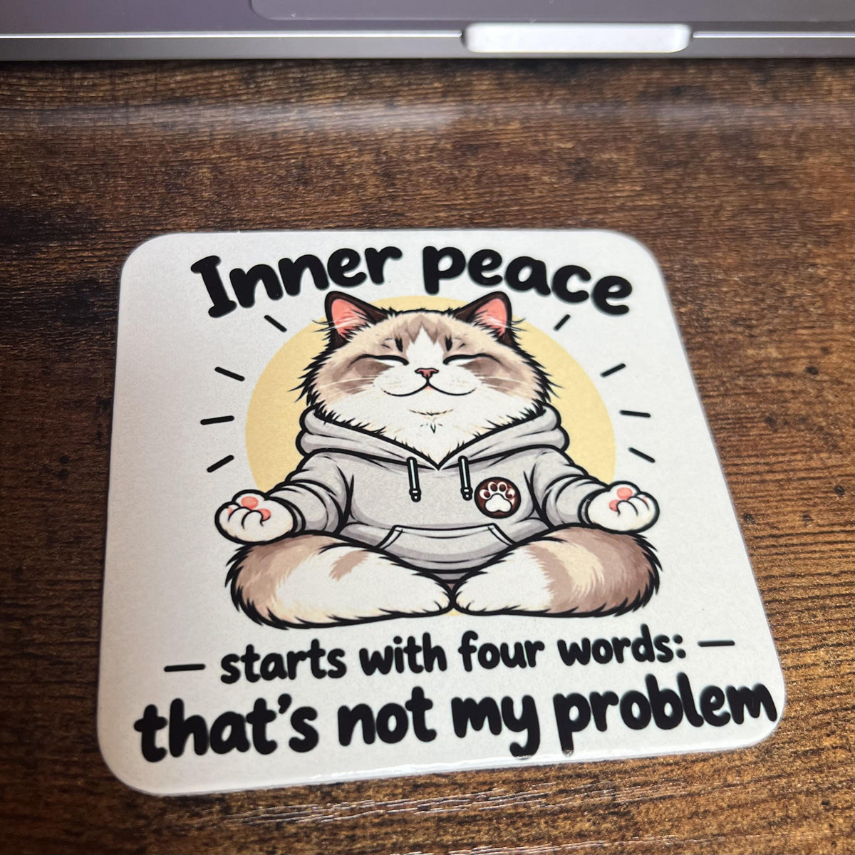 Korkový podtácek s kreslenou meditující ragdoll kočkou říkající "Inner peace starts with four words ..." na dřevěném stole před počítačem.