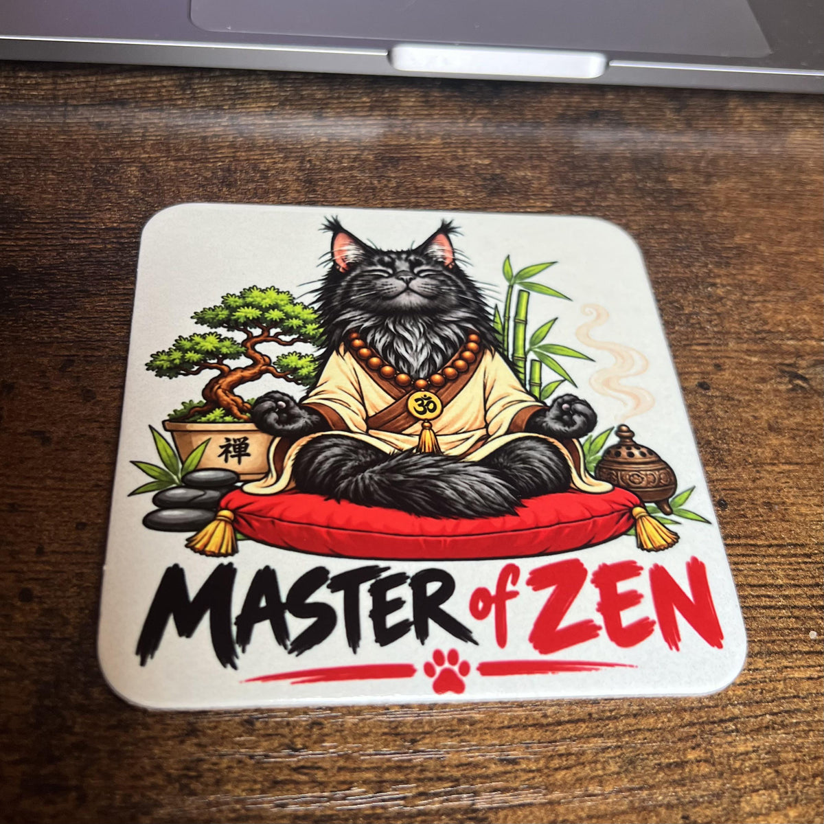 Korkový podtácek s kreslenou meditující černou mývalí kočkou říkající "Master of ZEN" na dřevěném stole před počítačem.